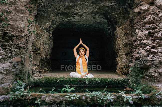 Woman with eyes closed meditating while sitting in lotus pose near stone cave — Stock Photo