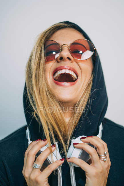 Jeune femme blonde en lunettes de soleil et sweat à capuche avec écouteurs riant sur fond blanc — Photo de stock