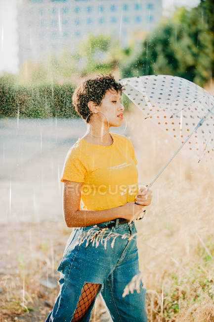 Junge Frau in stylischem Outfit schaut weg, während sie unter Wassertropfen mit Regenschirm läuft — Stockfoto