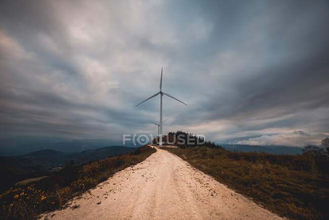 Fila de moinhos de vento modernos de pé no lado da estrada rural estreita no dia nublado — Fotografia de Stock