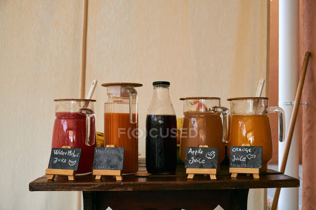 Diversi gusti di succhi colorati in brocche in piedi su scrivania in legno con frutta su sfondo parete beige — Foto stock