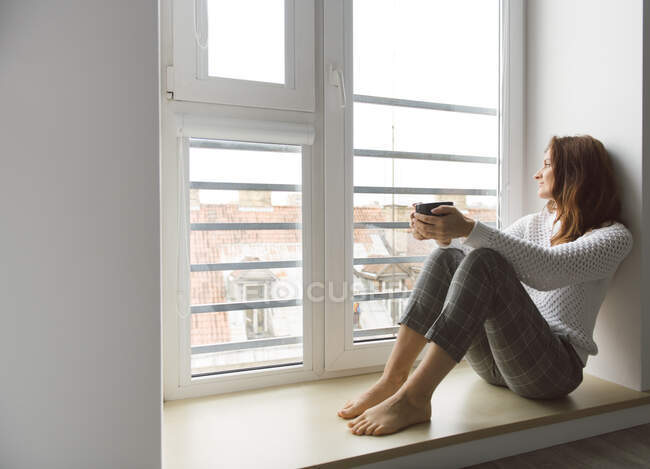 Сверху снимок привлекательной взрослой женщины в трикотажном свитере, держащей кружку горячего напитка и смотрящей на улицу, сидя на подоконнике в уютной комнате — стоковое фото