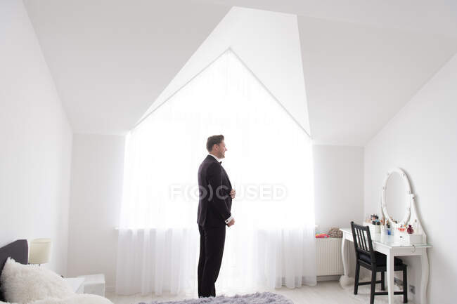 Side view of young handsome man standing in white bedroom and putting on black formal costume — Stock Photo