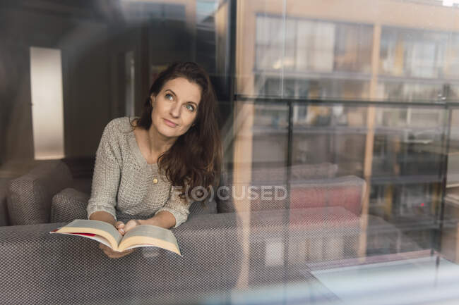 Красивая взрослая женщина в элегантном наряде улыбается и смотрит в сторону, лежа на удобном диване с интересной книгой — стоковое фото