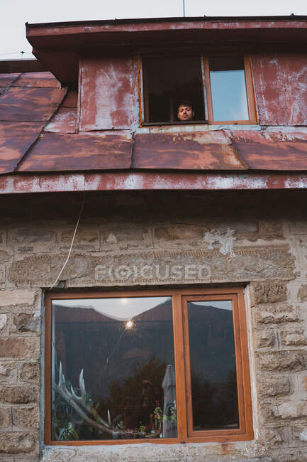Desde abajo plano de macho mirando por la ventana de la casa de campo resistido en pequeño asentamiento en Bulgaria, Balcanes - foto de stock