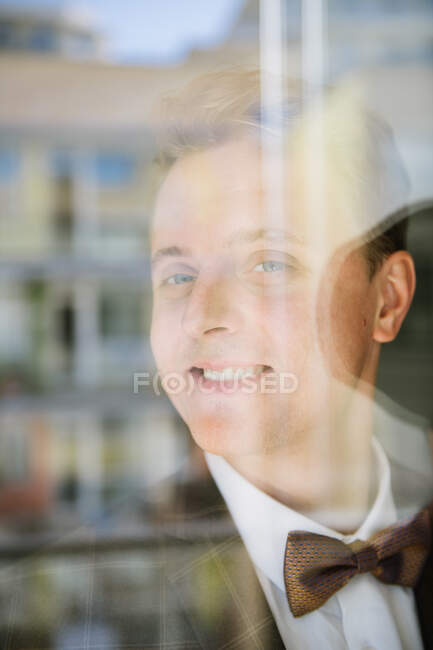 Schöner junger Mann in Anzug und Fliege lächelt und schaut in die Kamera, während er hinter Fensterglas steht — Stockfoto