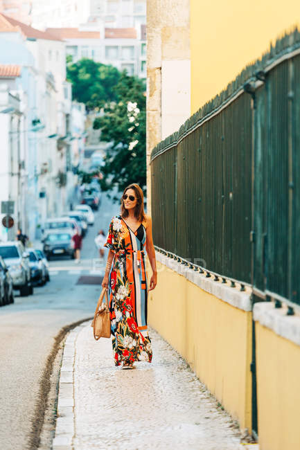 Элегантная женщина в длинном платье носит сумочку и ходит по улице под солнечным светом — стоковое фото