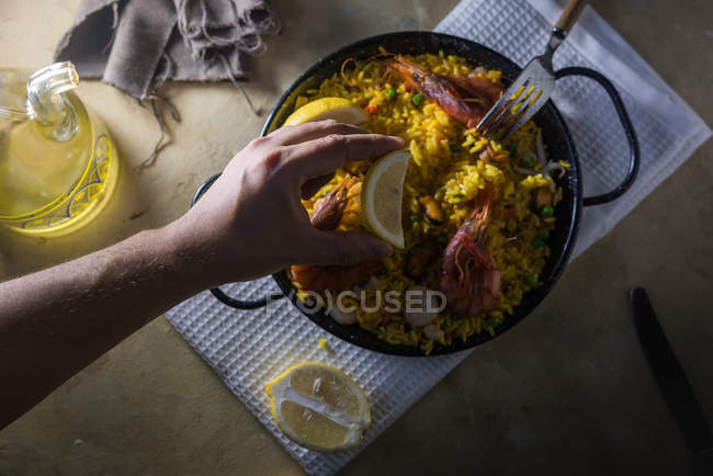 Mano humana exprimiendo limón sobre la tradicional paella marinera española con arroz, gambas, calamares y mejillones en sartén - foto de stock