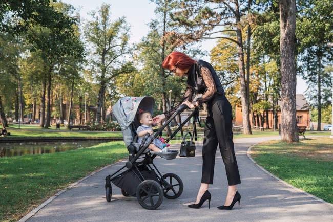 Элегантная молодая женщина, стоящая на аллее парка рядом с коляской с милой маленькой девочкой — стоковое фото