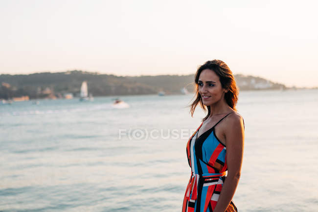 Content woman in colorful dress walking on cobblestone promenade at sunset against seascape — Stock Photo
