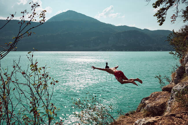 Back view of male with spreading hands jumping from scale in beautiful lake between mountains in sunny day in Austria — Foto stock