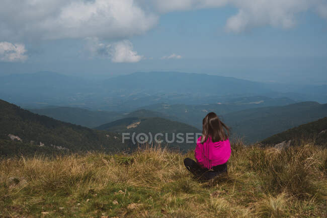 Back view of young woman in casual outfit sitting on top of grassy hill and admiring breathtaking view of beautiful nature on cloudy day in Bulgaria, Balkans - foto de stock