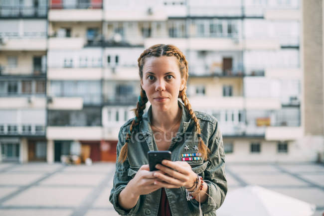 Retrato de menina de cabelos vermelhos com tranças usando telefone celular contra edifício residencial — Fotografia de Stock