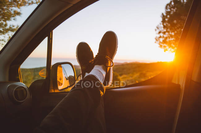 Las piernas del hombre anónimo acostado en la ventana del coche contra la vista de la magnífica campiña y el sol poniente brillante - foto de stock