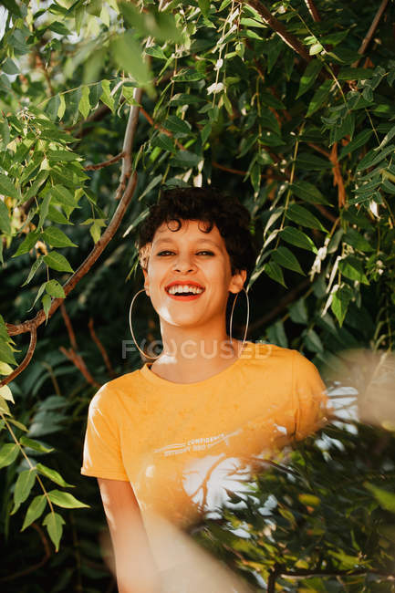 Портрет улыбающейся брюнетки с короткими волосами, стоящей в зеленой растительности с солнечным светом — стоковое фото