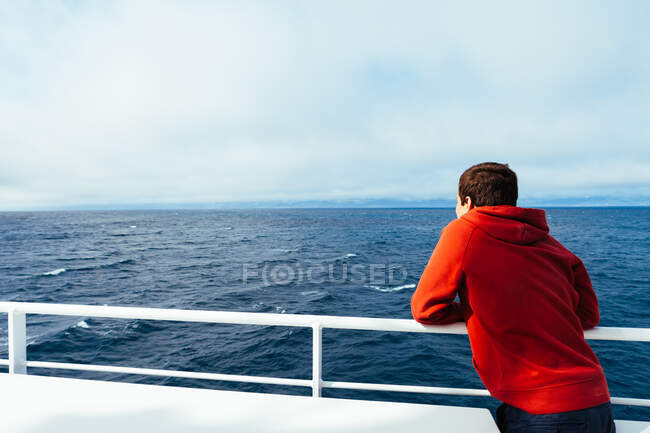 Вид сзади на ребенка в красной толстовке, стоящего на палубе корабля, смотрящего на бесконечные синие морские просторы — стоковое фото