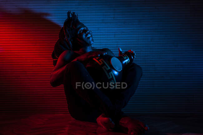 Young african rastafarian man enjoys rehearsing and plays udu, colored lighting red and blue — Stock Photo