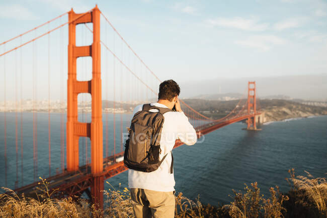Rückansicht eines nicht wiedererkennbaren Mannes mit Rucksack, der auf einer Klippe steht und majestätische goldene Torbrücke und Fluss an bewölkten Tagen in San Francisco fotografiert — Stockfoto