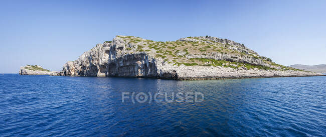 Steep rocky cliff covered with green grass and washed by blue clear water on background of cloudless sky — Stock Photo