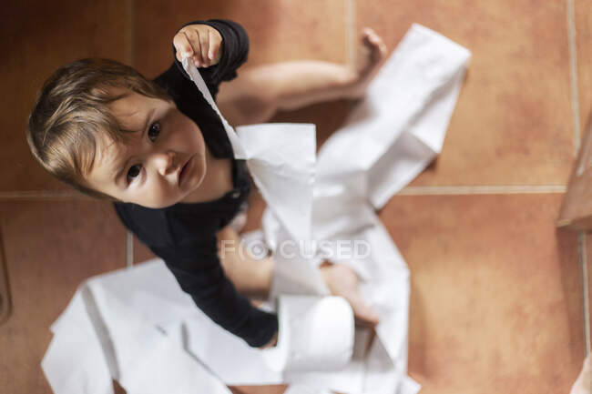 De arriba tiro de adorable bebé desenrollando papel higiénico sentado en el suelo mirando a la cámara - foto de stock