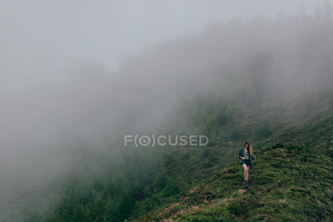 Frau geht auf hohem steilen Hügel, der mit grünem Gras bedeckt ist und über dem dichter Nebel liegt — Stockfoto
