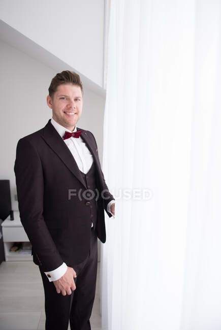 Vista lateral do jovem homem bonito em pé no quarto em traje formal preto e olhando através da janela atrás cortina branca — Fotografia de Stock