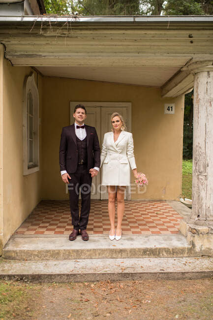 Hermoso hombre y mujer de moda en trajes de novia tomados de la mano y de pie en el porche de la casa vieja - foto de stock