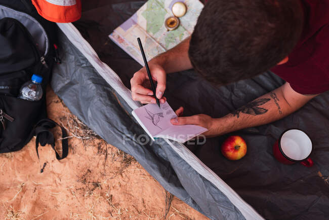 Von oben Aufnahme eines jungen Mannes, der im Zelt liegt und Skizzenbuch zeichnet, während er in der Wüste campt — Stockfoto