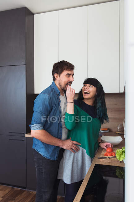 Vista lateral de la joven emocionada dando tomate cortado a novio alegre mientras se cocina en la cocina con estilo juntos - foto de stock