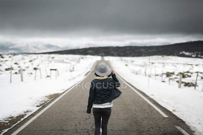 Вид на молодого человека в стильной одежде, идущего посреди асфальтовой дороги в пасмурный зимний день в красивой сельской местности — стоковое фото