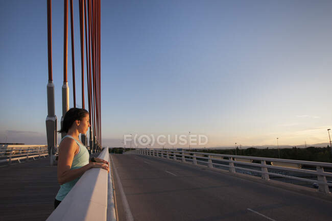Felice donna casuale appoggiata sulla recinzione del ponte e sorridente alla macchina fotografica in piena luce del sole — Foto stock