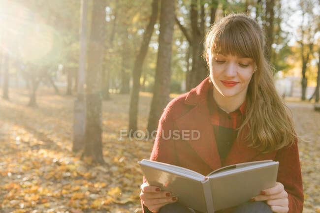 Attraktive junge Dame in rotem Mantel hält aufgeschlagenes Buch in der Hand und sitzt auf Sitz im Herbstwald — Stockfoto