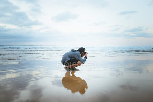Vue latérale d'un jeune homme anonyme assis sur du sable mouillé près d'une mer incroyable et prenant des photos par temps nuageux à Zarautz, Espagne — Photo de stock