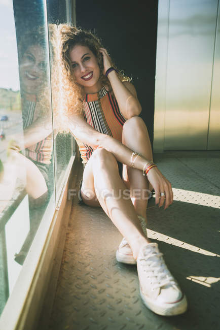 Sonriente joven mujer rizada sentado en el sol brillante cerca de la ventana - foto de stock