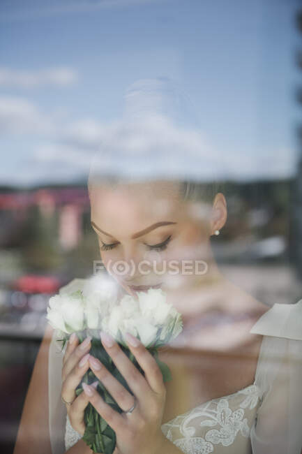 Linda noiva de vestido branco em pé na janela e cheirando monte de flores — Fotografia de Stock