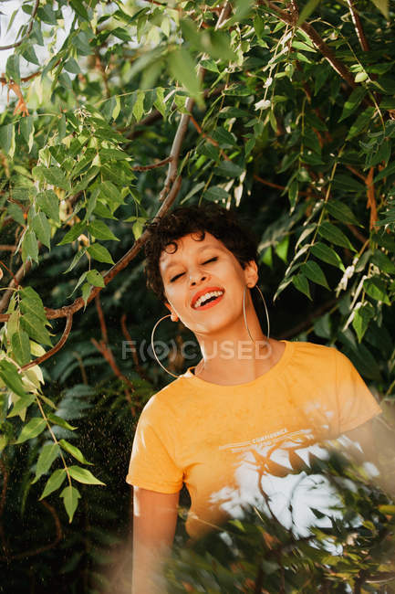Портрет усміхненої брюнетки з коротким волоссям, що стоїть у зеленій рослинності з сонячним світлом — стокове фото