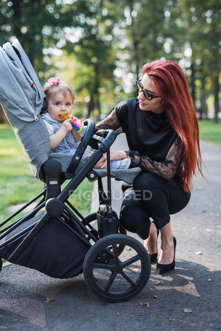 Jeune mère regardant bébé fille en poussette dans un parc ensoleillé — Photo de stock