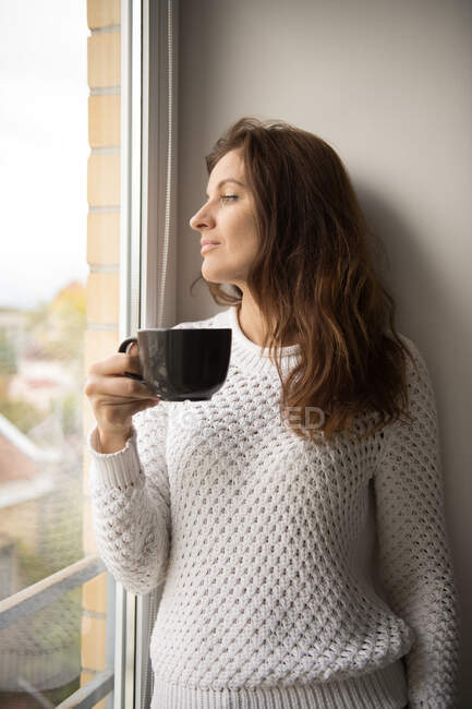 Beautiful adult female in knitted sweater smiling and looking away while holding cup of fresh hot beverage and leaning on wall — Stock Photo