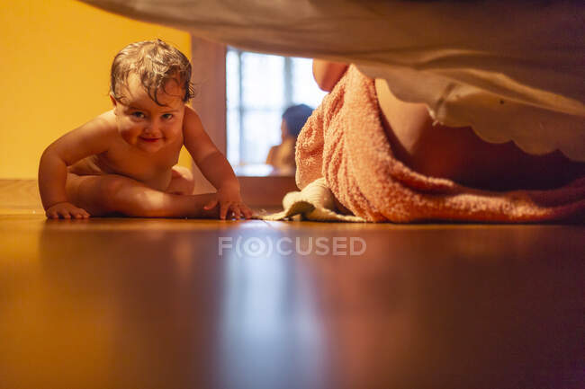 Criança bebê encantador molhado após o banho sentado no chão com a mãe perto de olhar sob a cama curiosamente — Fotografia de Stock
