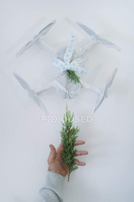 Main mâle tenant branche de sapin à côté de drone enveloppé comme cadeau de Noël sur fond blanc — Photo de stock