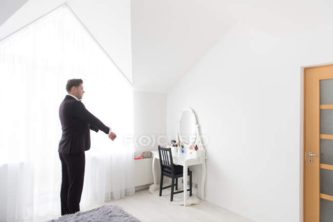 Vista lateral del joven hombre guapo de pie en el dormitorio blanco y ponerse el traje formal negro - foto de stock