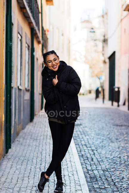Mulher de conteúdo bonito abraçando em casaco quente preto em pé na rua de paralelepípedos e rindo — Fotografia de Stock