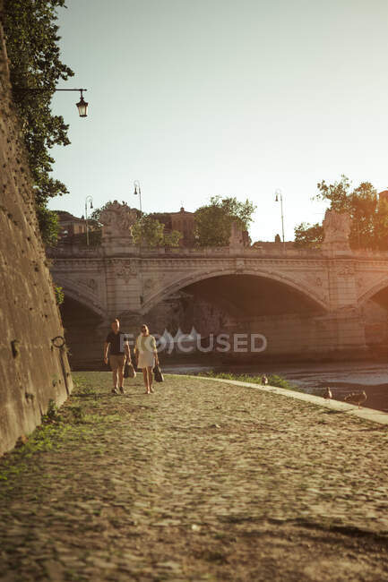 Jeune couple marchant sur la chaussée près de la rivière calme par une journée ensoleillée dans une Rome étonnante, Italie — Photo de stock