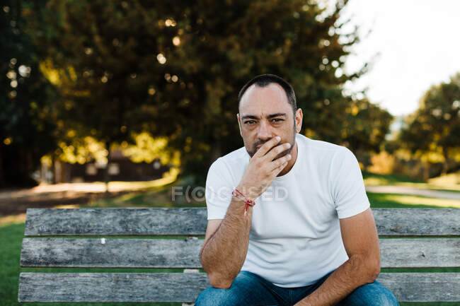 Привлекательный взрослый мужчина, прикрывающий рот, сидя на скамейке в парке в солнечный день — стоковое фото