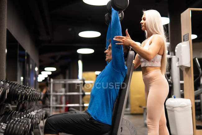 Femme aidant l'homme à faire de l'exercice avec des haltères dans la salle de gym — Photo de stock