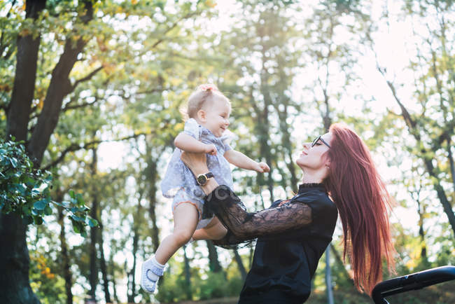 Pretty young woman with ginger hair holding baby girl while standing on blurred background of park trees on sunny day — Stock Photo