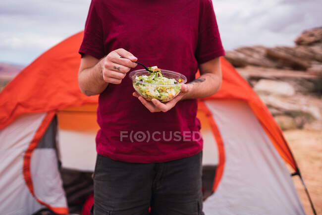 Crop man with bowl of fresh salad standing near modern tent on camping area in desert — Stock Photo