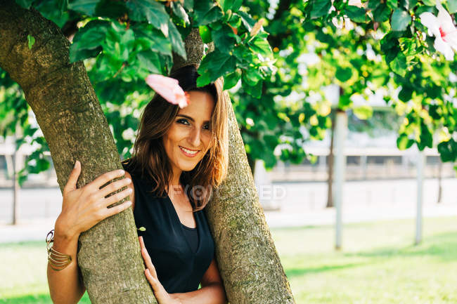 Cheerful stylish woman standing under tree embracing trunk and smiling at camera in park — Stock Photo