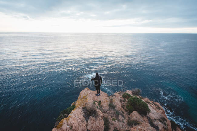 Назад вид неузнаваемого человека, стоящего на грубой скале и любующегося видом величественного се в пасмурный день — стоковое фото
