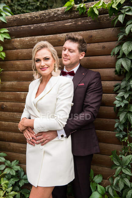 Elegante noiva adulta e noivo abraçando enquanto está de pé em ternos contra a parede de madeira e sorrindo para a câmera — Fotografia de Stock